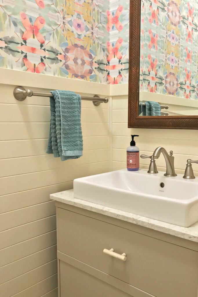 A small bathroom with a dresser vanity painted greige, white wainscoting, and colorful wallpaper.