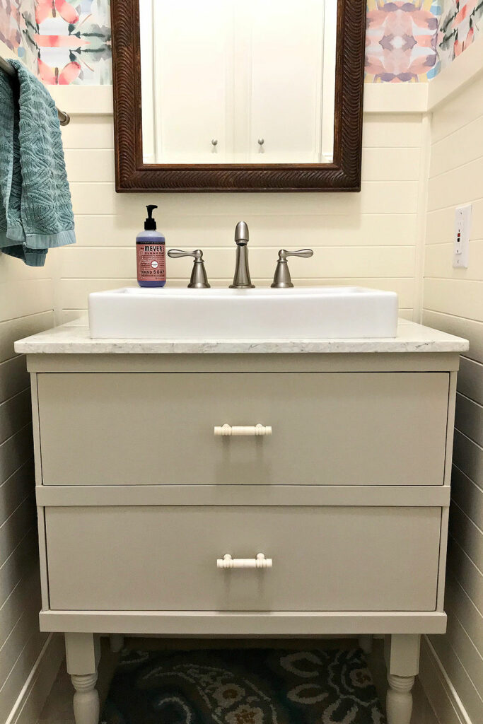 A greige vanity in a small bathroom with drawers and white drawer pulls. With a vessel sink on a quartz countertop.