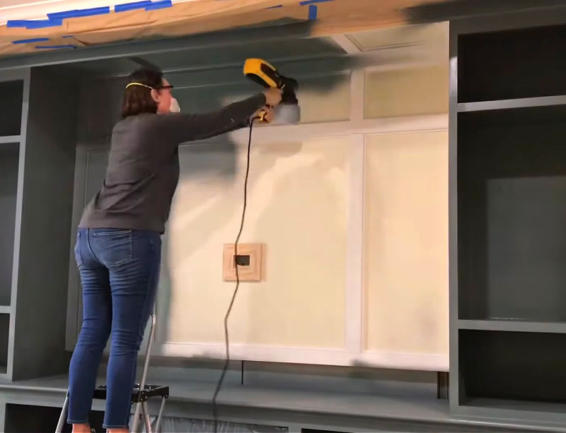 Image of someone painting built in bookshelves and cabinets with a paint sprayer.