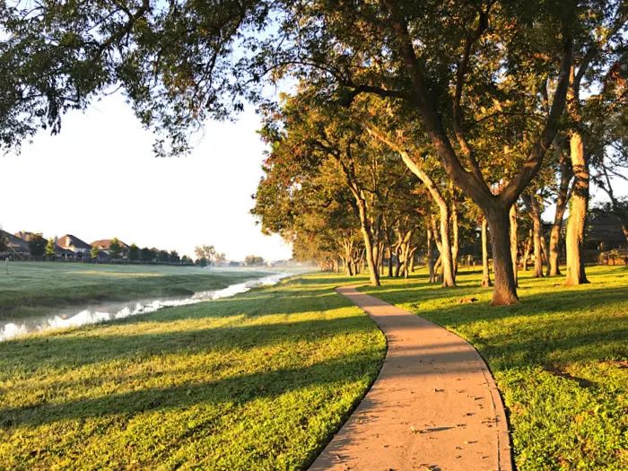 Walking trail with a sidewalk, trees, water, and grass.
