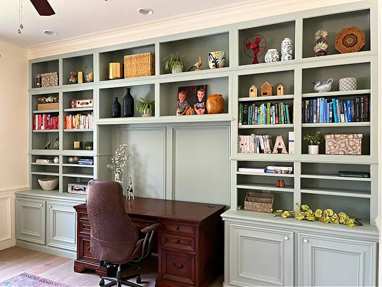 Home office with light sage green cabinet storage. Huge library style shelves in a home office with a stained wood desk.