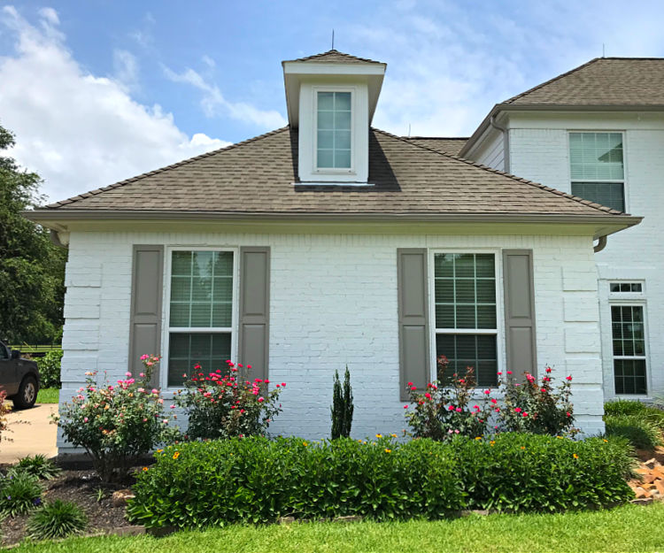 Side of a white brick house with grey-brown shutters and colorful landscaping.