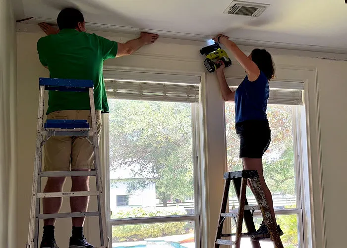 Image shows people installing three piece crown molding on a ceiling.